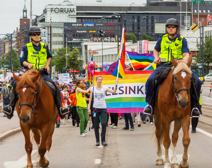 Kaksi ratsupoliisia ratsailla Pride-mielenosoituskulkueen kärjessä Mannerheimintiellä Helsingissä. Kulkueessa kannetaan sateenkaarilippuja.